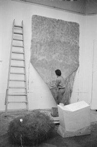 Installation view of <i>When Attitudes Become Form</i>. Keith Sonnier installing “Flocked Wall,” 1969. From left to right, works by Alan Sarret and Gary B. Kuehn. Kunsthalle Bern, Switzerland, 1969. Courtesy the Getty Research Institute, Los Angeles. Photo: Balthasar Burkhard. © J. Paul Getty Trust