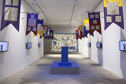 Installation view: Michael Smith, <em>Excuse me!?!...I'm looking for the “Fountain of Youth,”</em>, Greene Naftali Gallery, 2015. 12 screen printed cotton pennants, 6 channel video (looped), hand-blown glass bottle, wood, CO2 tank, speakers, haute lisse, hand woven tapestry, 100% wool. Courtesy of the artist and Greene Naftali, New York.
