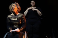 Alice Sheppard seated in her wheelchair holds a book and speaks directly to the audience. Candace Davider is over Alice's left shoulder making the ASL sign of emotional struggle, two open clawed hands going up with one hand and down with the other on the body several times. Both of their faces have soft expressions of internal conflict. Photo: Mengwen Cao. 