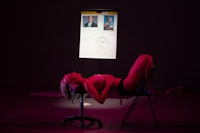 Audre Wirtanen performing <em>DX ME FIX ME</em>, Gibney Dance, 2020. Photo: Scott Shaw.<br><em>Audre, a thin white hypermobile femme, lies on her back, balancing on a medical office stool and folding chair. Her shins press against the back of the chair while her hands rest on her ribcage. Pink light bathes her entire body while a blurry easel collaged with pictures of doctors, a drawing, and text is lit bright white in the background. The easel says THE MEN WHO FUCKED ME. Audre is in her underwear with ECG electrodes stuck to her breasts like medical pasties. Her expression is heavy and silent.</em>