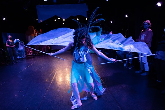 <em>TERRITORY: The Island Remembers</em> at Gibney Center. Photo: Scott Shaw.<br><br> <strong>Alt text:</strong> In a shiny blue feathered costume with sheer fabric wings dotted with origami cranes, Raha Behnam dances in tribute to the mythical Simurgh, a Persian bird representing collectivity.