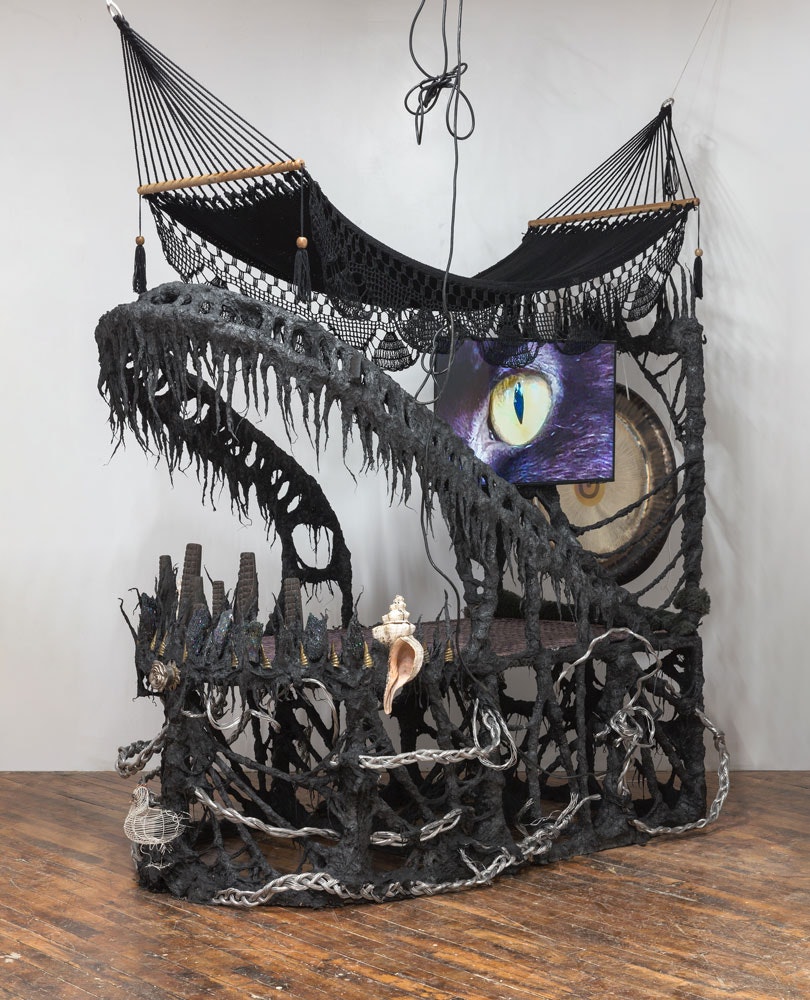 Guadalupe Maravilla, <em>Disease Thrower #0</em>, 2022. Gong, hammock, LCD TV, ceremonial ash, pyrite crystals, volcanic rock, steel, wood, cotton, glue mixture, plastic, loofah, and objects collected from a ritual of retracing the artist's original migration route, 96 x 68 x 98 inches. Courtesy PPOW Gallery.