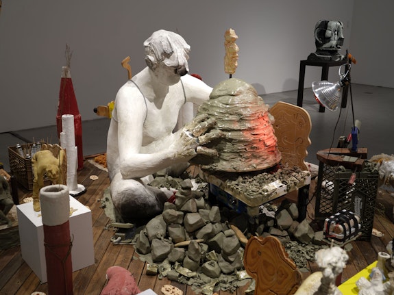 Nicole Eisenman, <em>Maker’s Muck</em>, 2022. Coney Island Boardwalk (Brazilian Ipe), plaster, bronze, silicone, unfired clay, fired clay, expanding foam, burlap, wire, raw wool, sneakers, magic smooth, magic sculpt, resin, bamboo skewers, tin foil, plaster bandages, plywood, bass wood, Aqua-Resin, fiberglass, seashell, styrofoam, oil paint, wax, carpet fringe, cardboard, cement, steel pipe, steel rod, aluminum paint tubes, vinyl stickers, granite, telescopic extension pole, sand, fabric, crocheted rug, marbles, miniature rocking chair, grass, woven rush stool, Formica, Le Beau Touché plasteline, canola oil, corn, Valvoline motor oil, sponge, reflective fabric, nylon cord, charcoal, Shimpo Whisper pottery wheel, Saran wrap, coated wire, recording device, AM/FM Radio, wood clamp, spring clamp, expansion clamp, heat lamp bulb, clip light, glass bottles, Plexiglas, dried flowers, SLS plastic, dried plastic, milk crate, canvas, twine, copper tube, elastic cord, steel I-beam, MDF, iron object, 103 1/4 x 120 x 155 1/4 inches. © Nicole Eisenman. Courtesy the artist and Hauser & Wirth. Photo: Thomas Barratt.